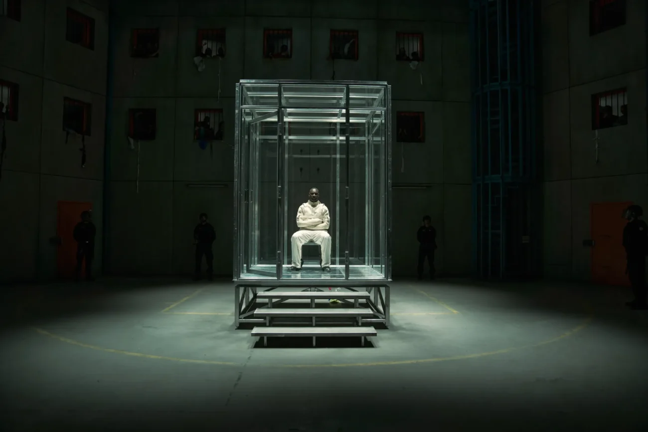 "A person in a white outfit sits alone inside a glass cell at the center of a dimly lit, industrial room with concrete walls. Several shadowy figures stand in the background, emphasizing a tense, isolated prison environment."
