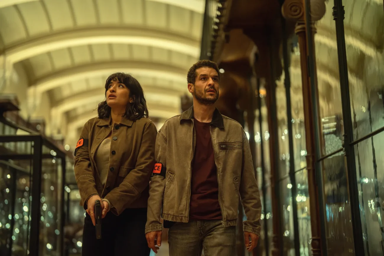 "Two people wearing jackets with armbands walk through a dimly lit, ornate hallway lined with glass cases. One holds a gun, both look alert and cautious, as if investigating or searching for something. The setting feels tense and mysterious."