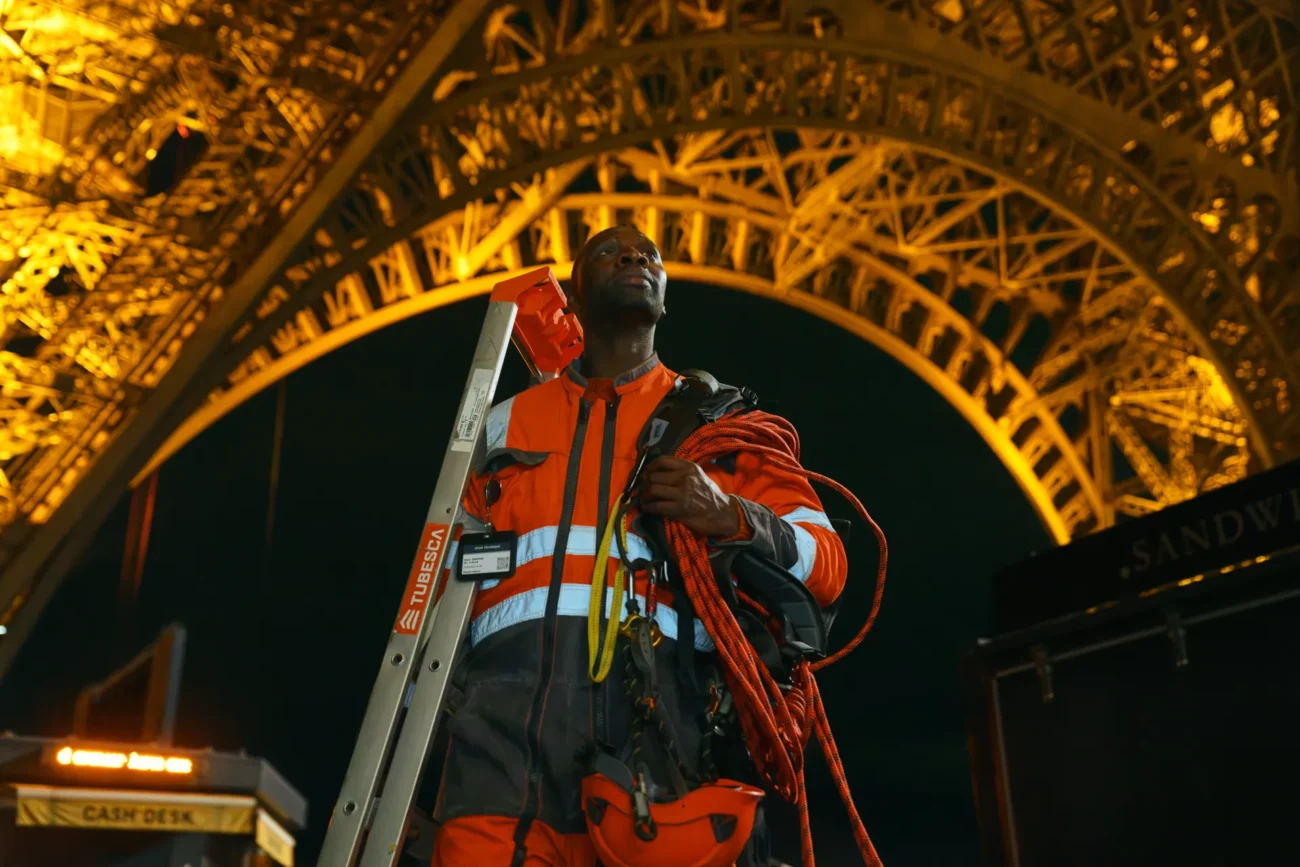 "A worker in orange safety gear stands under the illuminated Eiffel Tower at night, holding a ladder and rope, with dark sky and structural metal details visible above."