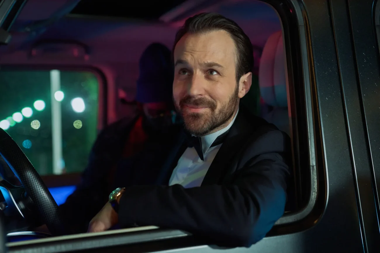 "Man in a suit sitting in the driver's seat of a car at night, looking out the window and smiling, with city lights visible outside and a passenger in the background."