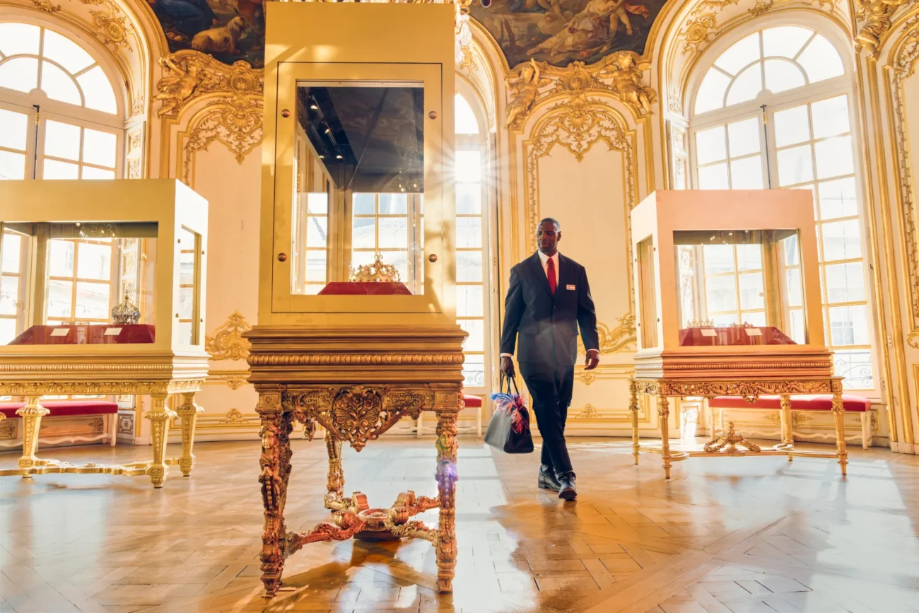 "A security guard walks through an ornate, sunlit museum gallery with large windows, golden decorative walls, and glass display cases showcasing artifacts on elegant tables."