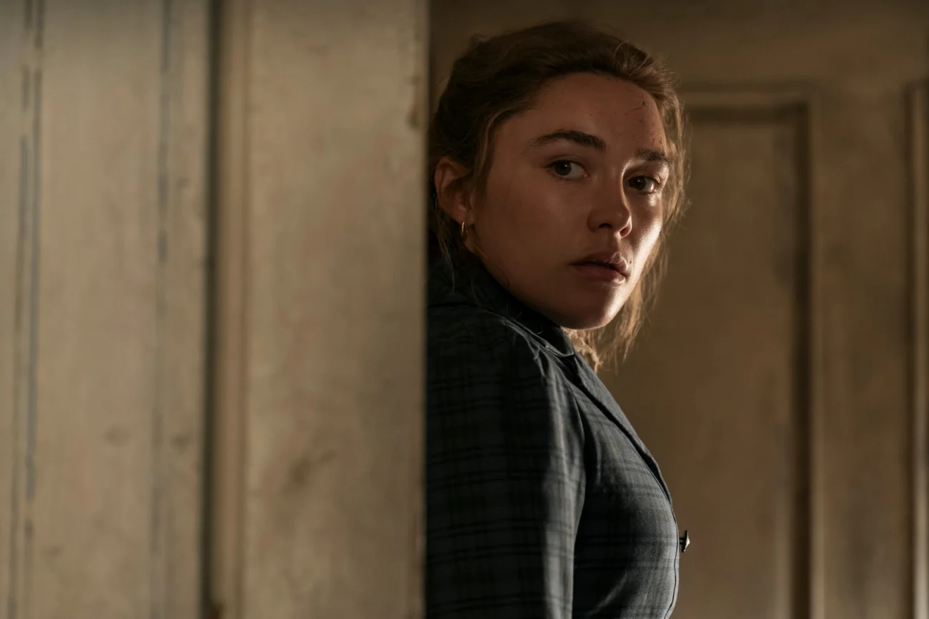 "A woman in period clothing stands partially hidden in a dimly lit room, looking over her shoulder toward the camera with a serious expression. Worn wooden panels and neutral tones create an old, somber environment."